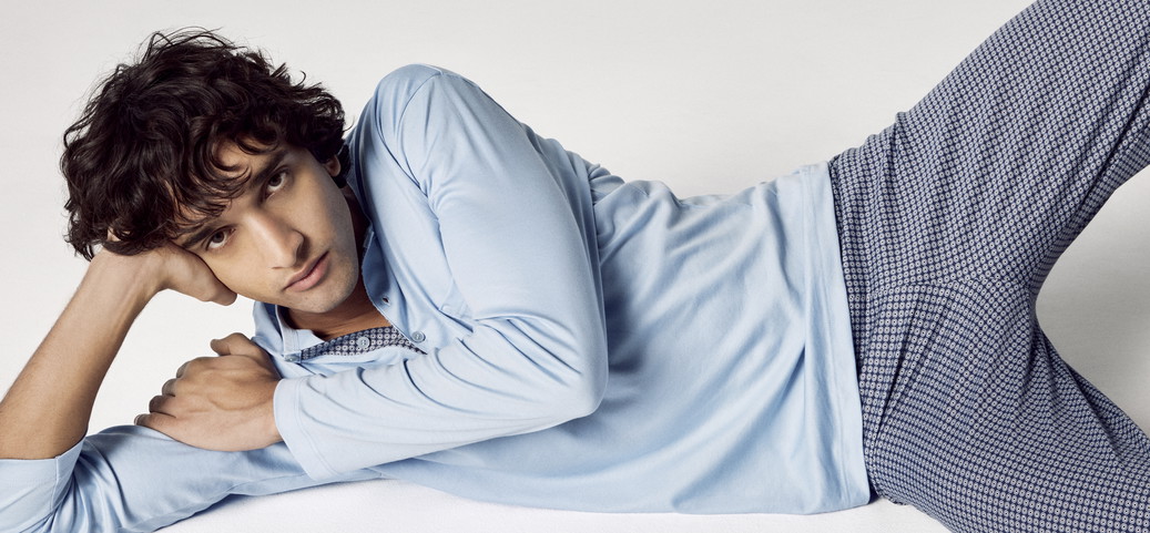 Homme allongé sur le côté sur une surface claire, portant une chemise bleu clair à manches longues et un pantalon bleu à motifs