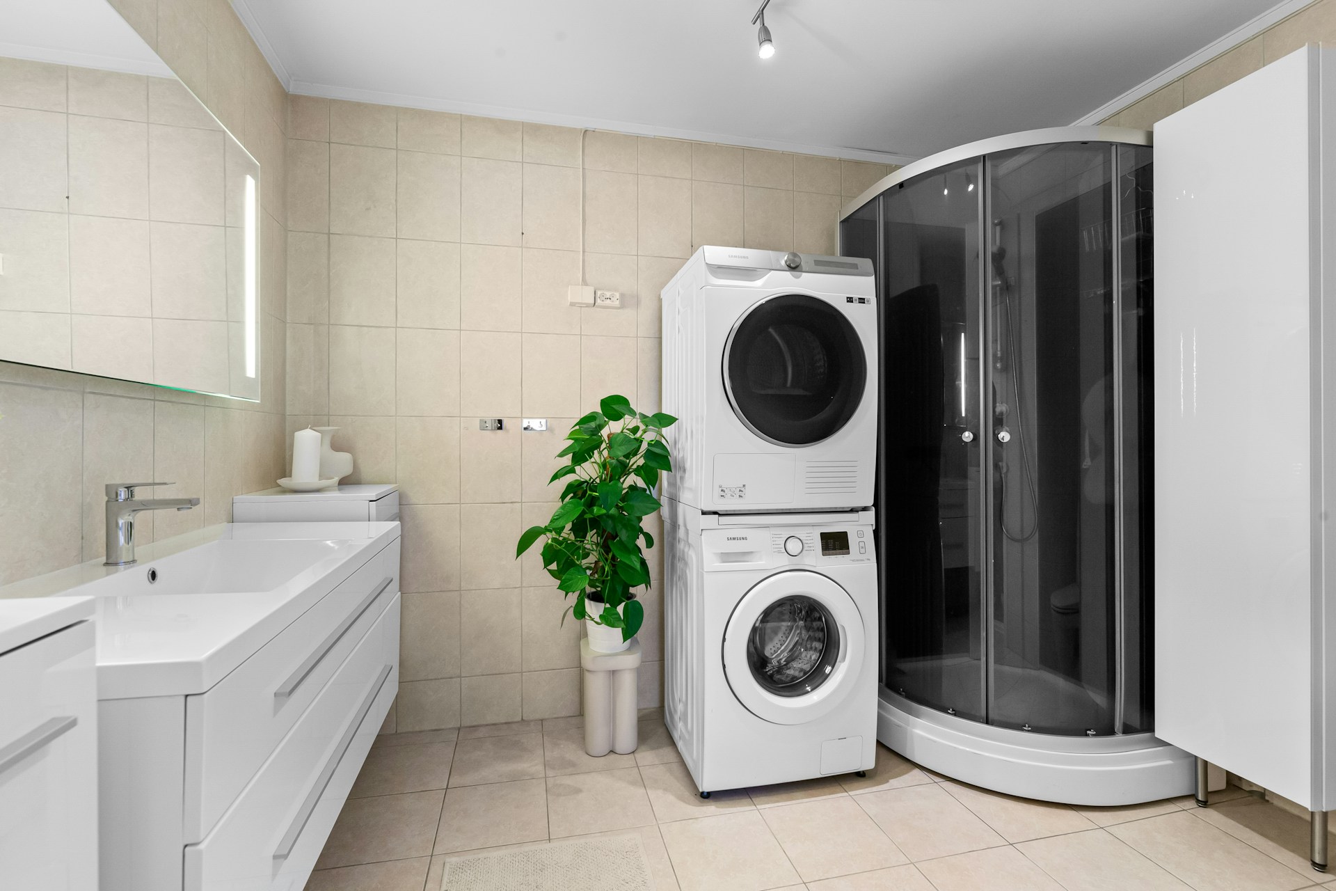 Modernes Badezimmer mit Waschbecken, beleuchtetem Spiegel, übereinander gestapelter Waschmaschine und Trockner, Glasdusche und grossem Pflanzengefäss auf hellen Fliesen