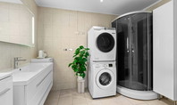 Salle de bain moderne avec lavabo, miroir éclairé, lave-linge et sèche-linge empilés, douche en verre et grand pot de plante sur carrelage clair