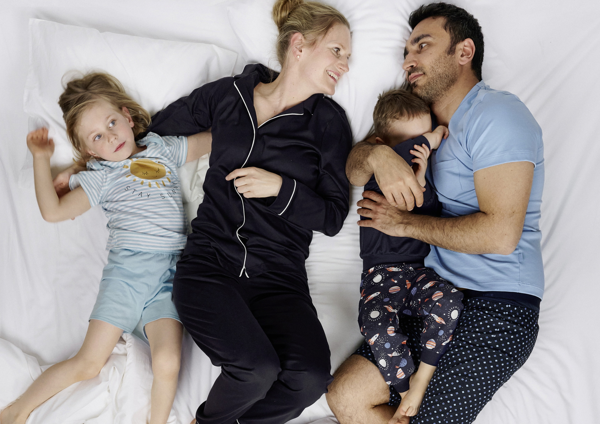 Familie liegt gemeinsam auf weissem Bett; zwei Erwachsene und zwei Kinder tragen Nachtwäsche von CALIDA, entspannte Schlafszene als Kontext dafür, warum man im Pyjama schlafen sollte