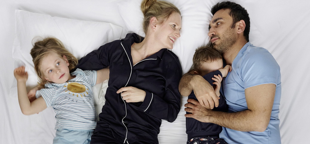 Familie liegt gemeinsam auf weissem Bett; zwei Erwachsene und zwei Kinder tragen Nachtwäsche von CALIDA, entspannte Schlafszene als Kontext dafür, warum man im Pyjama schlafen sollte