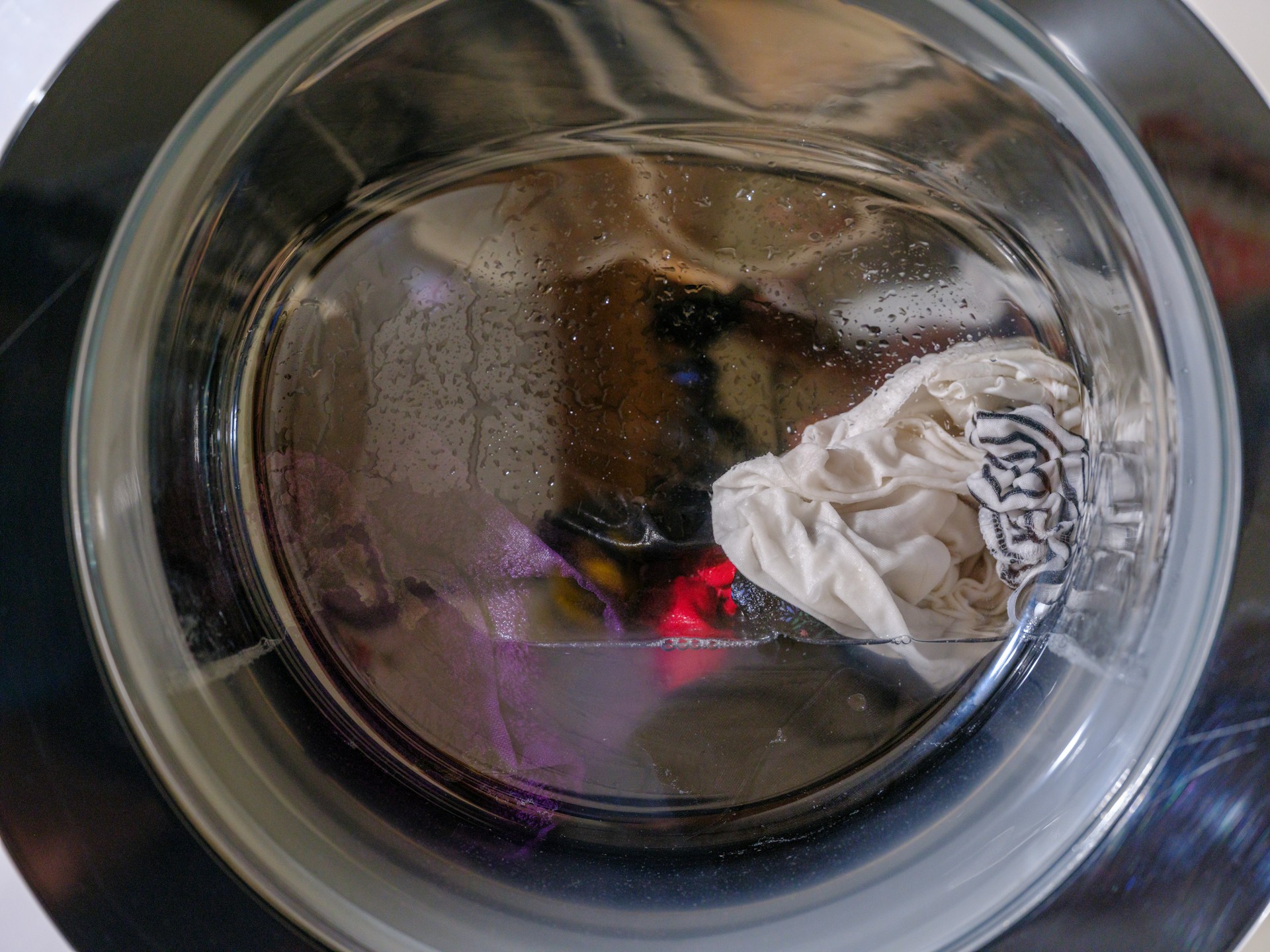 Nahaufnahme einer Waschmaschinentrommel, in der sich nasse, weiße und farbige Kleidung befindet. Es sind Wassertropfen auf der Innenseite der Trommel sichtbar.