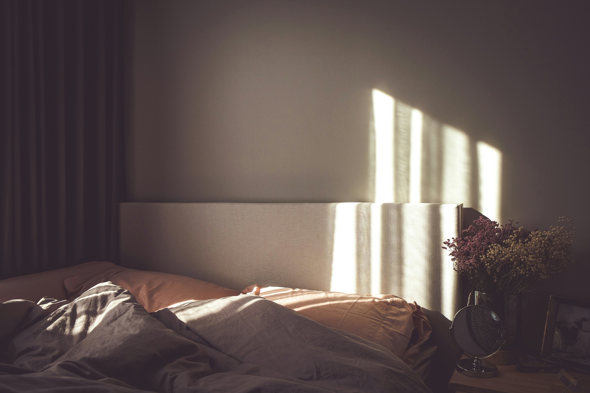L'image montre un lit avec deux oreillers et des couvertures, éclairé par un rayon de soleil. À côté du lit se trouve une table de nuit avec un miroir, un tableau et des fleurs. La pièce semble confortable et accueillante.
