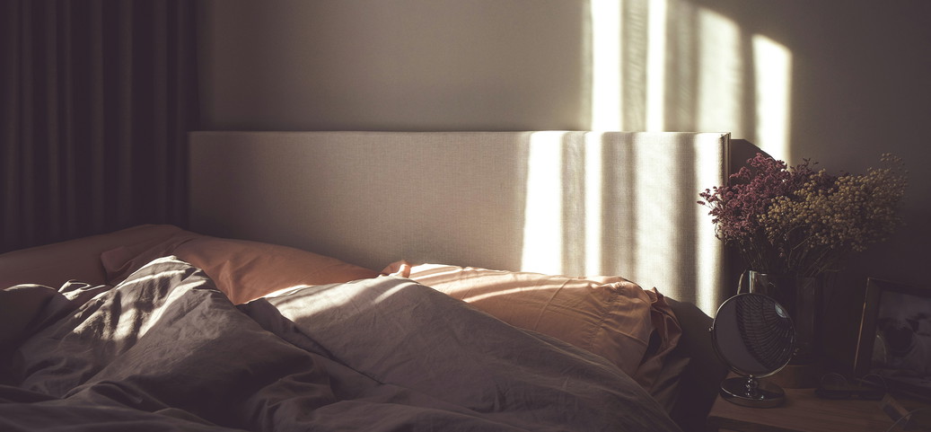 L'image montre un lit avec deux oreillers et des couvertures, éclairé par un rayon de soleil. À côté du lit se trouve une table de nuit avec un miroir, un tableau et des fleurs. La pièce semble confortable et accueillante.