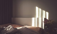 L'image montre un lit avec deux oreillers et des couvertures, éclairé par un rayon de soleil. À côté du lit se trouve une table de nuit avec un miroir, un tableau et des fleurs. La pièce semble confortable et accueillante.