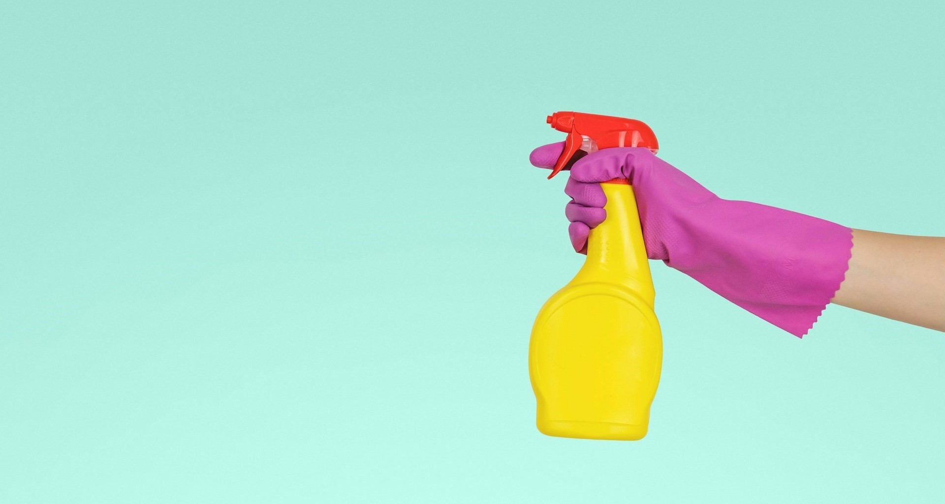 Une main portant un gant rose tient un vaporisateur jaune à bouchon rouge sur un fond turquoise.
