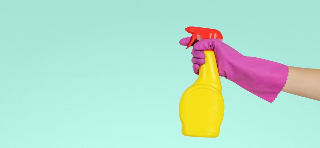 A hand with a pink glove holds a yellow spray bottle with a red lid in front of a turquoise-coloured background.