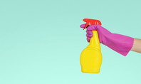Une main portant un gant rose tient un vaporisateur jaune à bouchon rouge sur un fond turquoise.