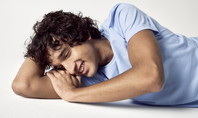 L'image montre un homme souriant aux cheveux bouclés, allongé sur le côté sur un fond blanc et portant un T-shirt bleu clair de CALIDA. Il a les yeux fermés, ce qui doit symboliser le rire pendant le sommeil.