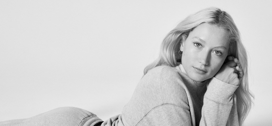 Une femme est allongée sur le sol et s'appuie sur ses coudes. L'image est en noir et blanc. Elle porte un sweat-shirt gris.