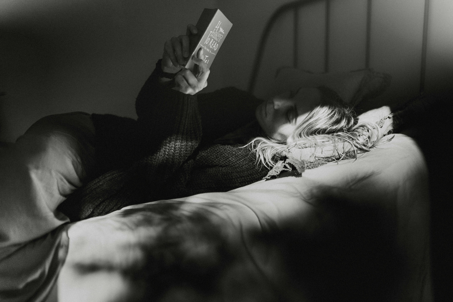 Person liegt entspannt in einem Bett und liest ein Buch im schwachen Licht, um zur Ruhe zu kommen und besser einschlafen zu können.