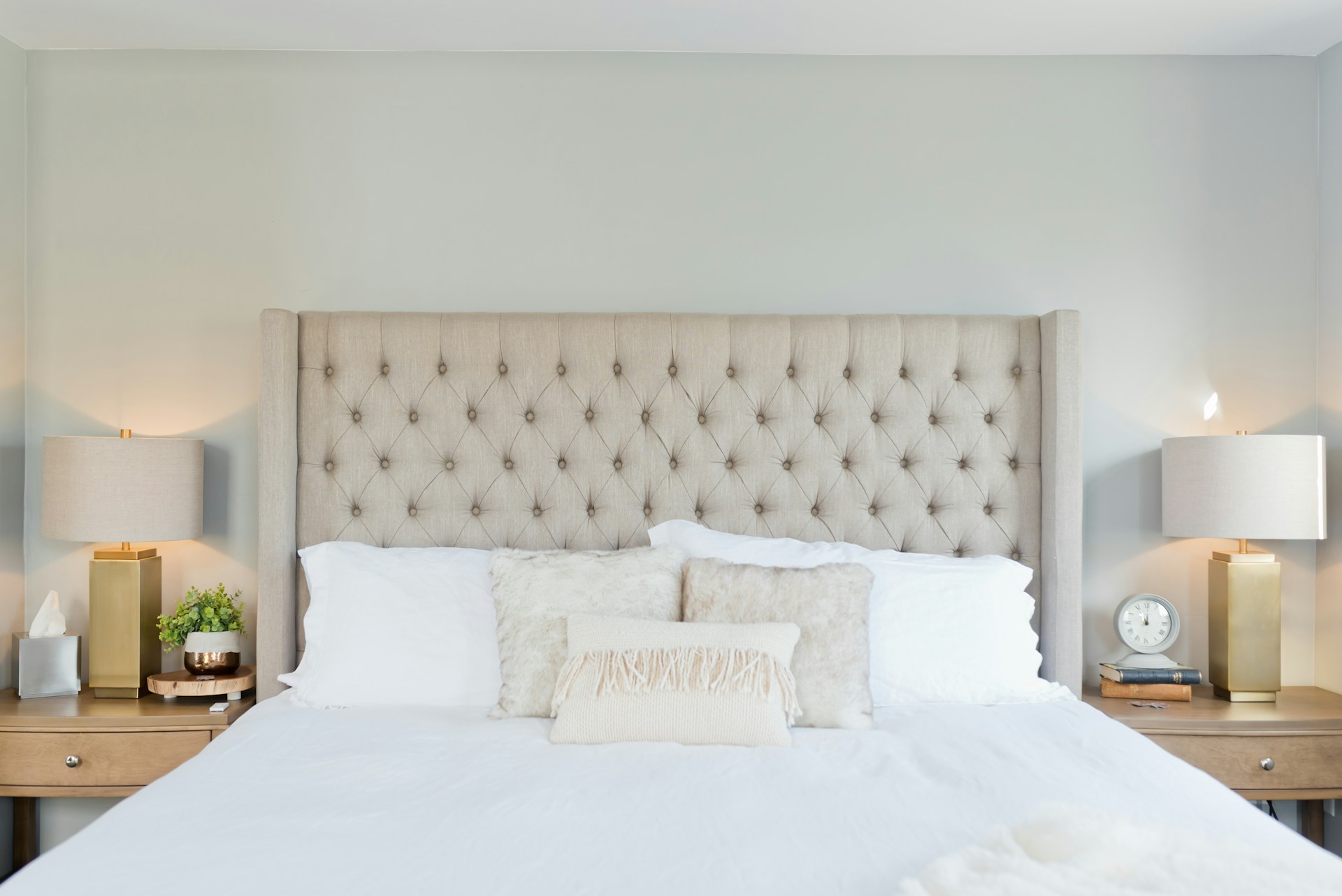 Helles Schlafzimmer mit beigem, gepolstertem Kopfteil, zwei Nachttischen, goldenen Lampen und weißer Bettwäsche – Symbol für trockene Luft im Schlafzimmer.