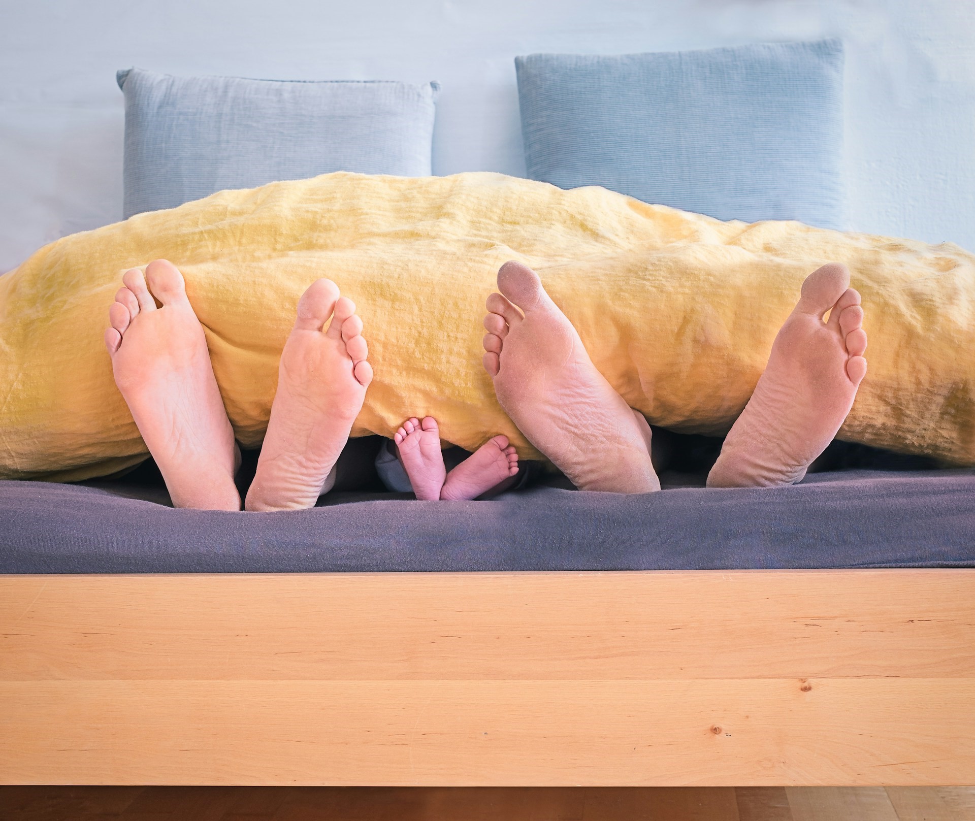 Eltern und Baby beim Co-Sleeping, ihre Füße schauen gemeinsam unter der Bettdecke hervor.