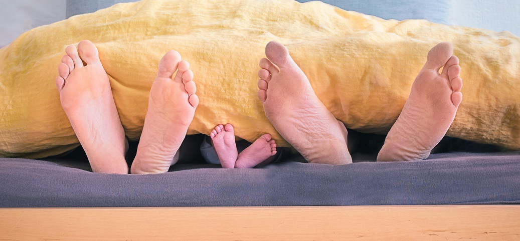 Eltern und Baby beim Co-Sleeping, ihre Füße schauen gemeinsam unter der Bettdecke hervor.