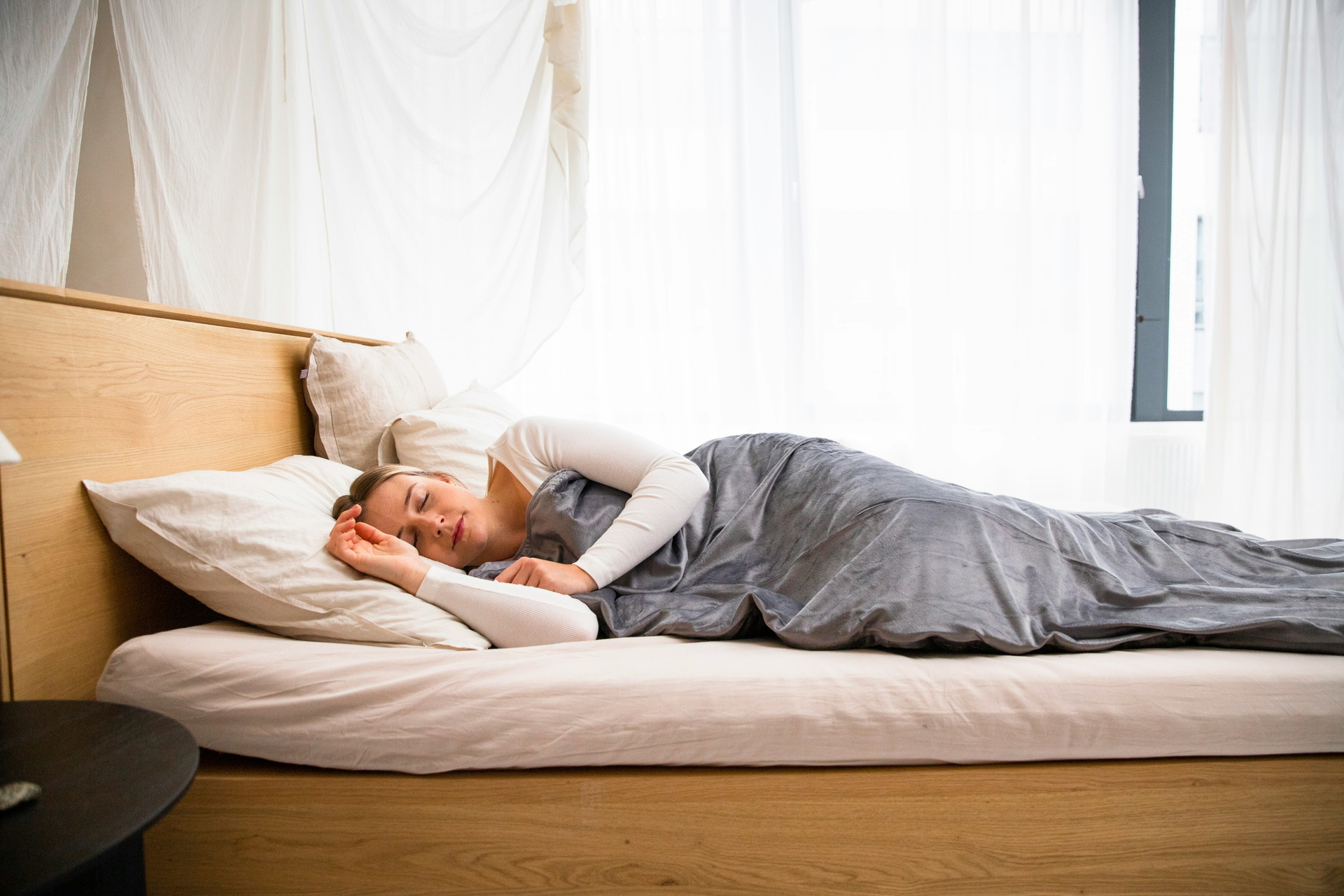  Auf dem Bild liegt eine Frau seitlich im Bett, ihre Augen sind geschlossen und ihr Gesicht zeigt Anzeichen von Entspannung. Die Frau ist von einer grauen Decke bedeckt, die sanft über ihren Körper drapiert ist. Unter ihrem Kopf liegt ein weißes Kissen, das eine weiche Unterstützung bietet. Die Szene strahlt Ruhe und Gelassenheit aus, während die Frau in einem Moment der Ruhe und des Friedens verweilt.