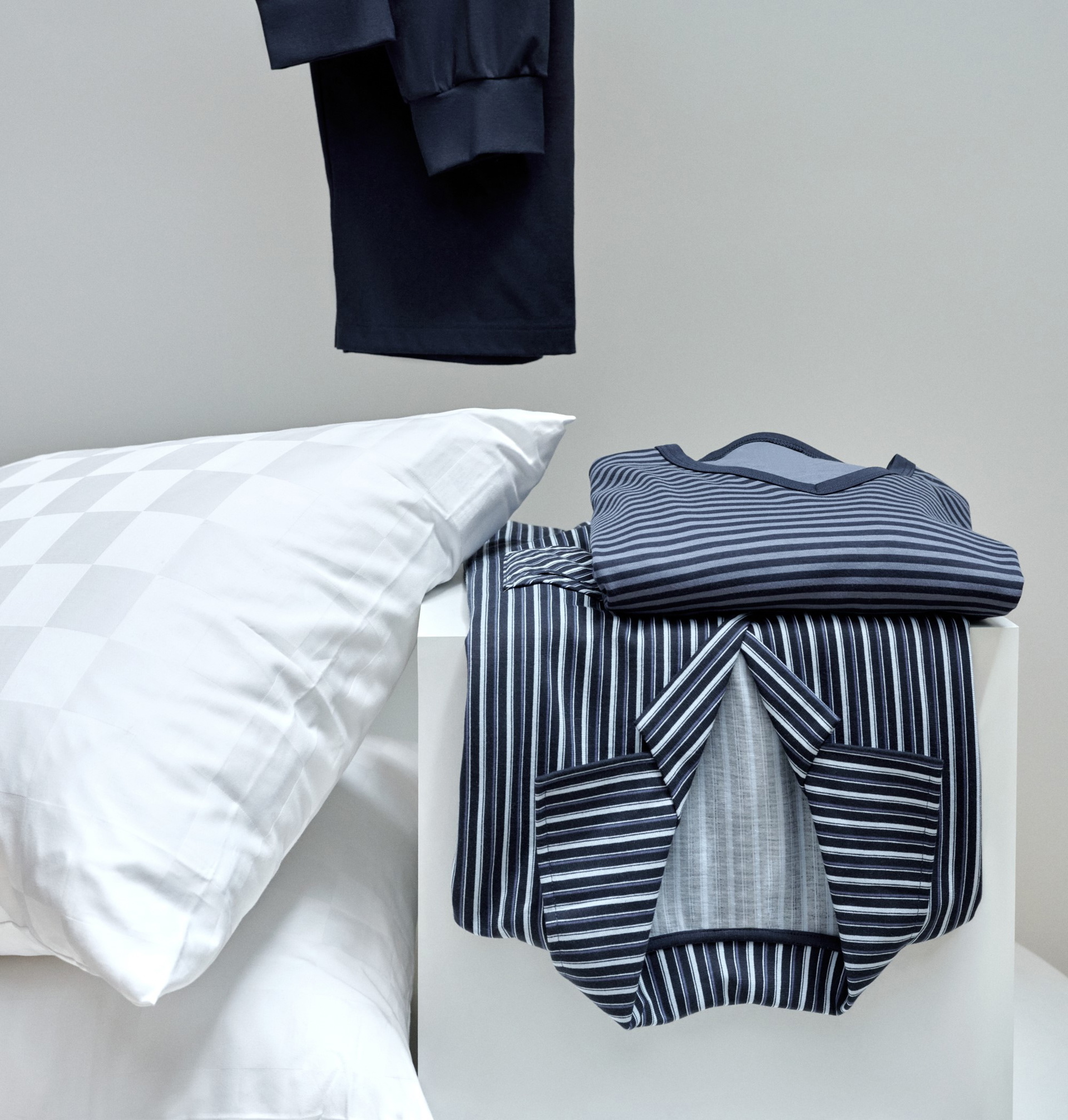 Pyjama à rayures plié dans les tons bleus et gris sur une table blanche à côté d'un coussin blanc avec un motif à carreaux discret. Un vêtement noir est suspendu à l'arrière-plan.
