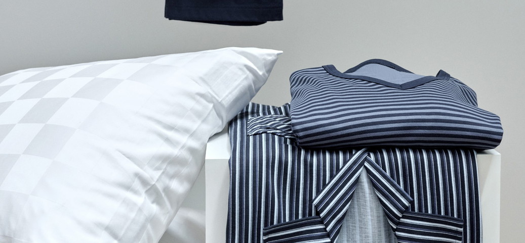 Pyjama à rayures plié dans les tons bleus et gris sur une table blanche à côté d'un coussin blanc avec un motif à carreaux discret. Un vêtement noir est suspendu à l'arrière-plan.