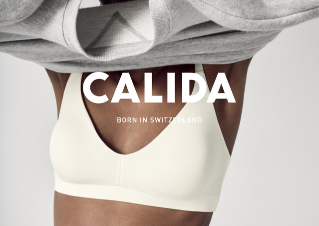 La femme retire un pull gris et porte un soutien-gorge crème en dessous. Grand logo 'CALIDA' avec 'Born in Switzerland' en dessous.