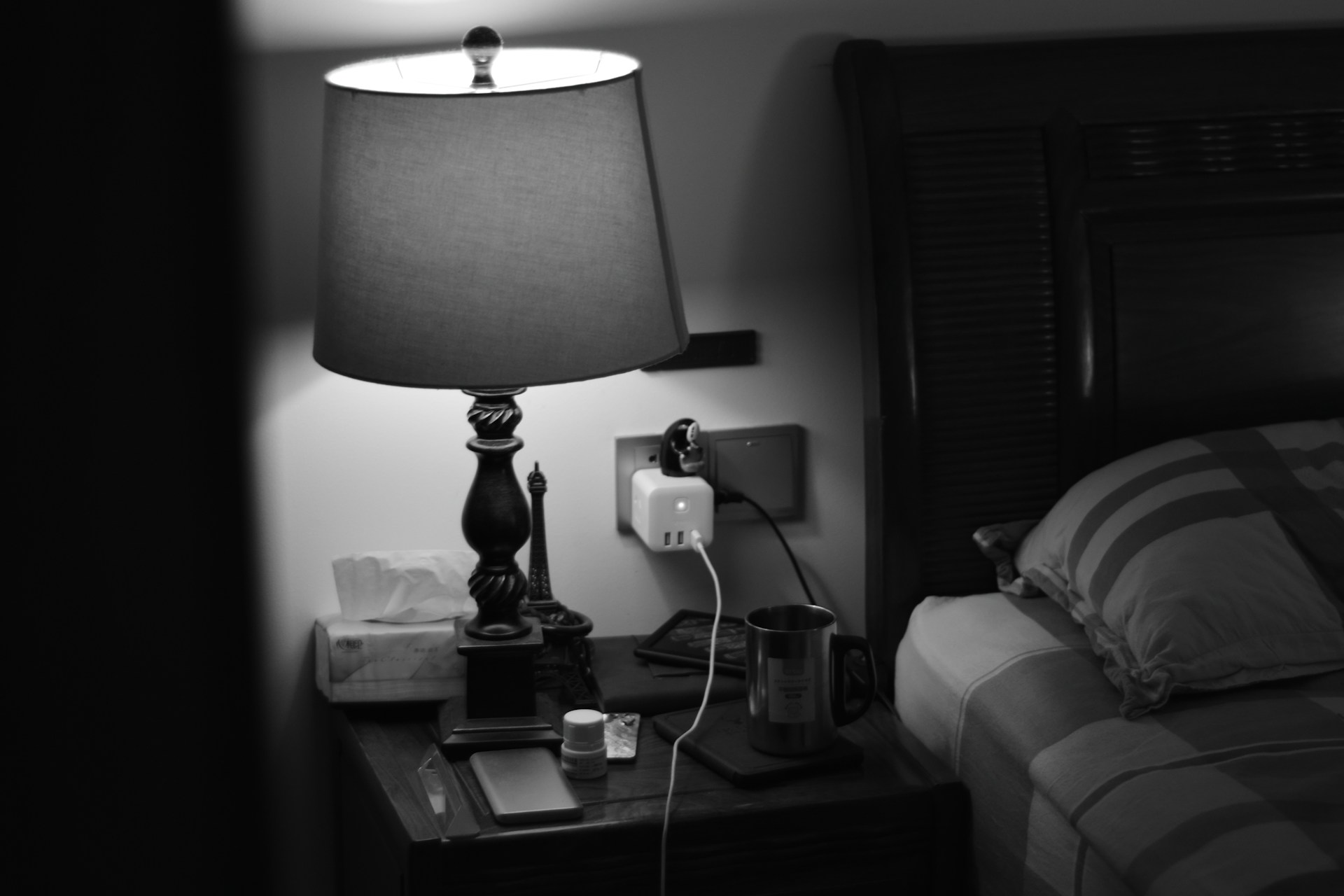 Comodino con lampada, caricabatterie nella presa, tazza di metallo e smartphone accanto a un letto con cuscino.