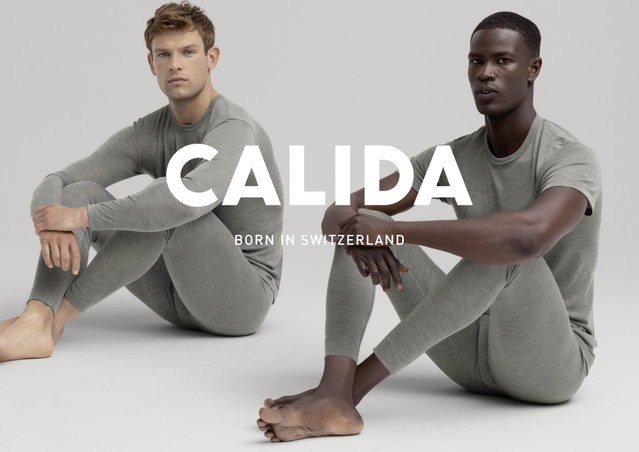 Zwei Männer sitzen auf dem Boden in graugrüner Loungewear, einer trägt ein langärmeliges Oberteil, der andere ein T-Shirt. Beide schauen ernst in die Kamera. Großes Logo CALIDA mit dem Slogan Born in Switzerland im Vordergrund.