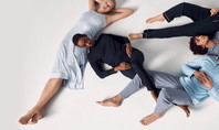 Sur l'image, cinq personnes sont allongées en cercle, chacune avec les yeux fermés. Leurs têtes reposent chacune sur le ventre de l'autre. Toutes portent des vêtements de nuit de Calida et sont allongées sur le dos. La scène dégage une impression de calme et de sécurité, tandis que les personnes se détendent dans leurs positions de sommeil confortables.