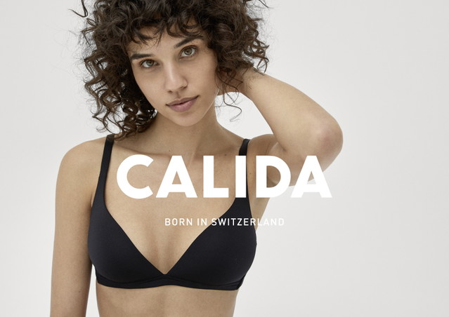 Une femme assise sur un sol blanc porte un soutien-gorge triangle noir et une culotte de la série ECO SENSE par CALIDA.
