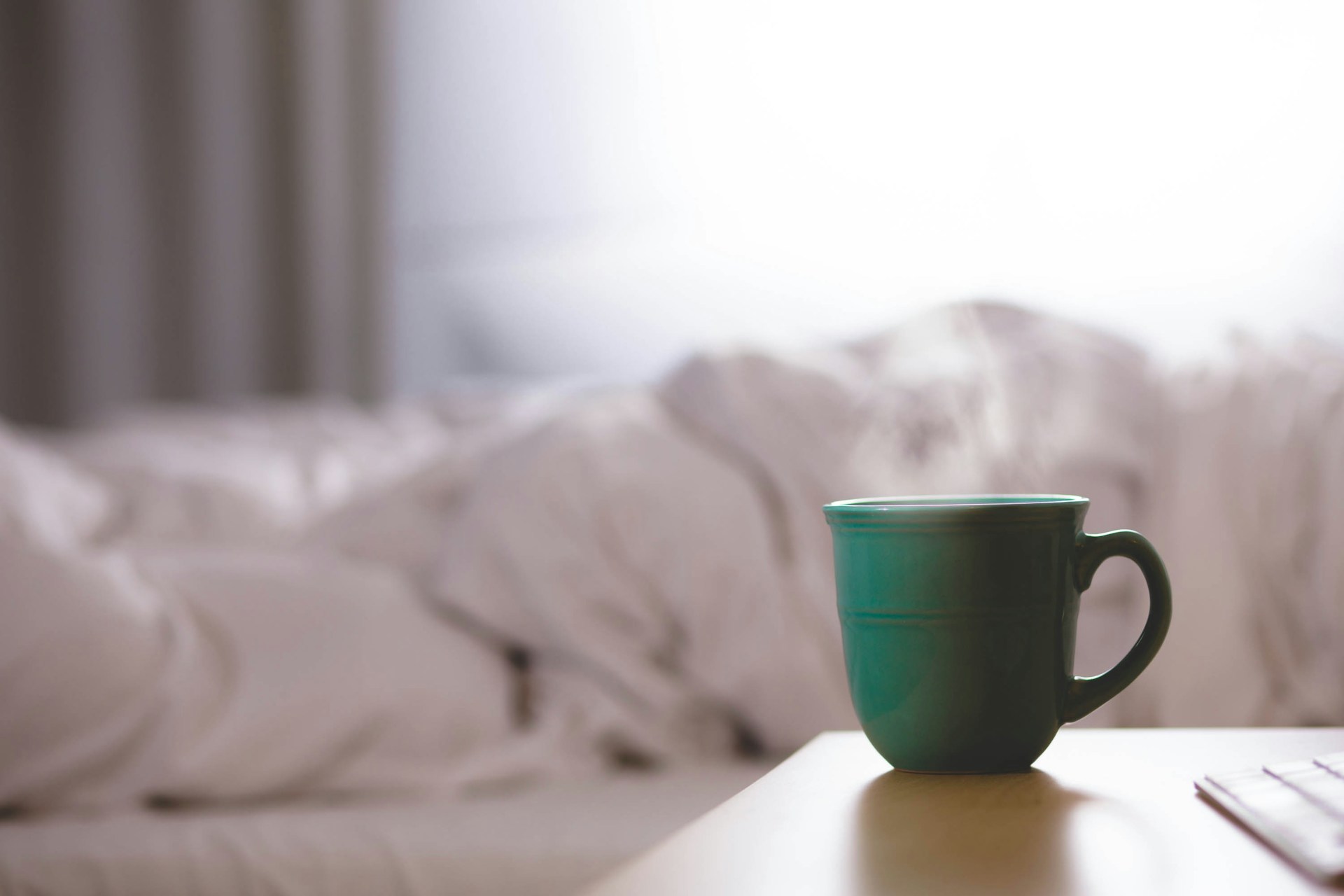 Une tasse verte remplie de café fumant est posée sur une table de nuit devant un lit défait, symbole du café avant de dormir.