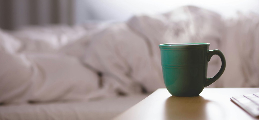 Une tasse verte remplie de café fumant est posée sur une table de nuit devant un lit défait, symbole du café avant de dormir.