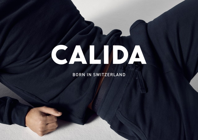 La personne est allongée et détendue dans un loungewear sombre, le haut légèrement relevé laisse entrevoir la ceinture du pantalon. Grand logo 'CALIDA' avec 'Born in Switzerland' en dessous.