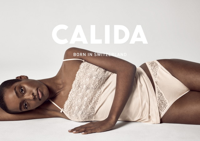 Dunkelhäutige Frau liegt seitlich auf einem hellen Untergrund, trägt ein zartrosafarbenes Spitzentop mit schmalen Trägern und eine passende Unterhose mit Spitzenverzierung. Großes Logo CALIDA mit dem Slogan Born in Switzerland im Vordergrund.