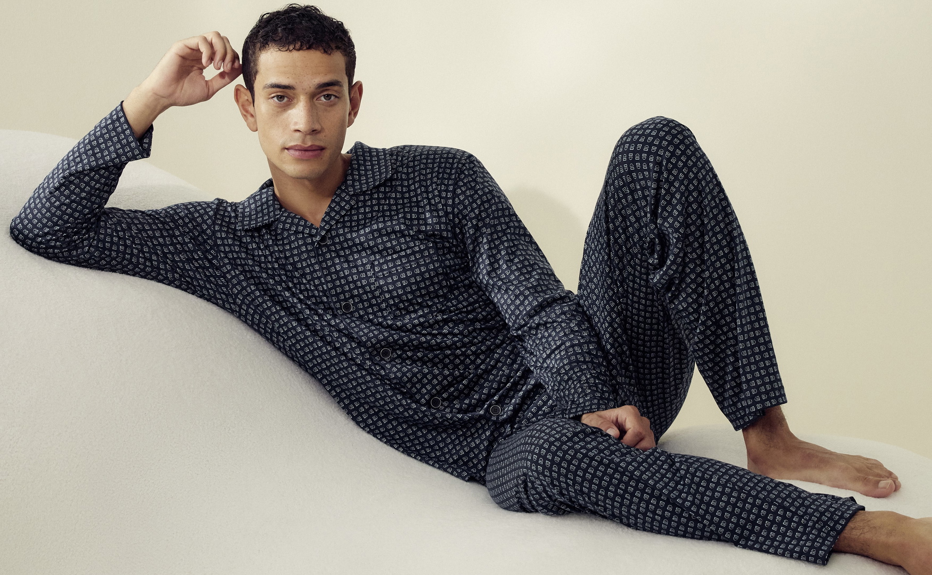 Man sitting on a soft surface. His right arm is bent so that he is touching his hair with his fingers. He is wearing Calida nightwear from the 'Relax Selected' range  