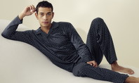 Man sitting on a soft surface. His right arm is bent so that he is touching his hair with his fingers. He is wearing Calida nightwear from the 'Relax Selected' range