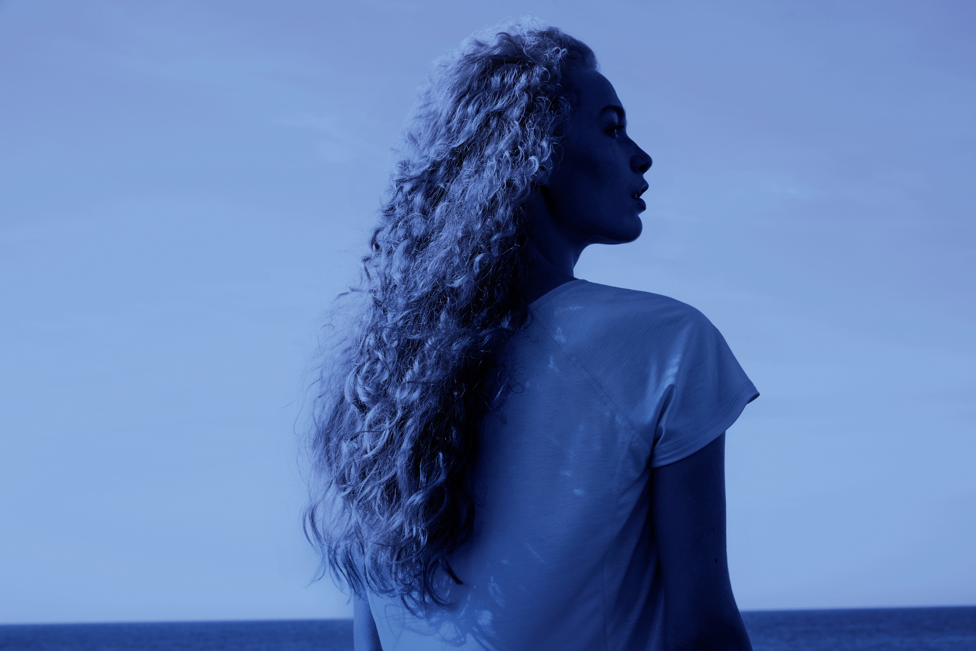 Une femme aux cheveux longs et bouclés portant un vêtement de nuit CALIDA léger, regardant la mer dans un cadre de nuit bleu profond et frais.