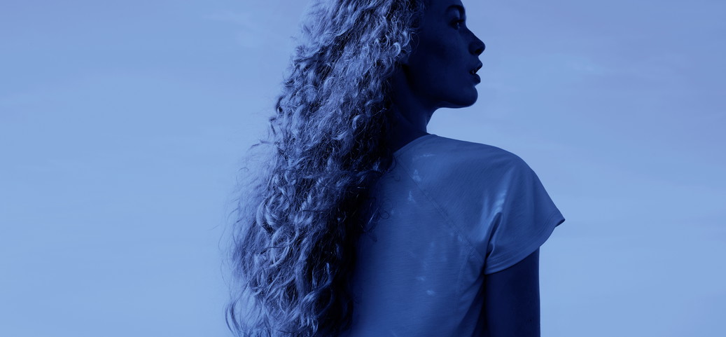 Une femme aux cheveux longs et bouclés portant un vêtement de nuit CALIDA léger, regardant la mer dans un cadre de nuit bleu profond et frais.
