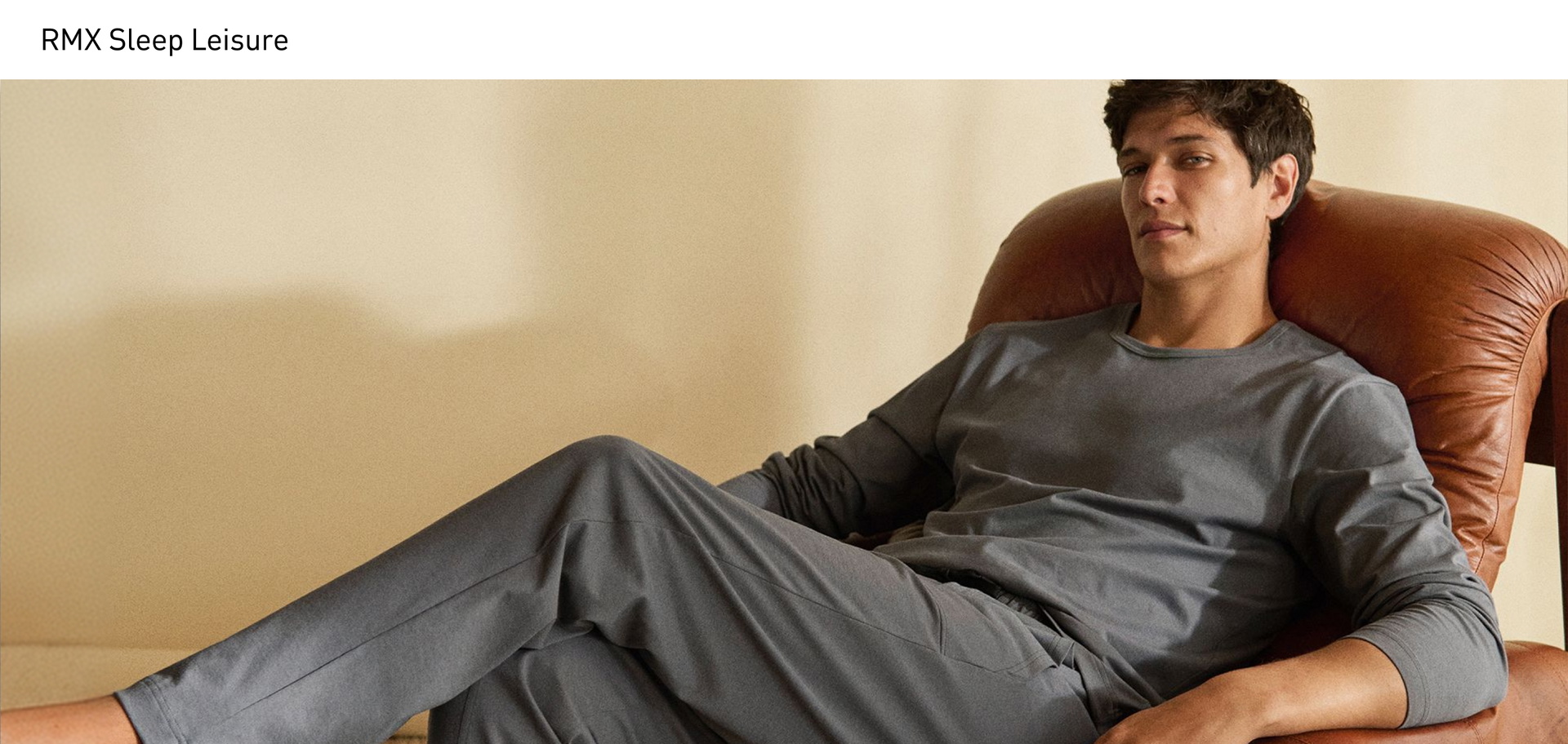 A man is sitting comfortably in an armchair, wearing a grey long-sleeved shirt with matching pyjama trousers featuring side pockets and a drawstring from the RMX Sleep Leisure series.