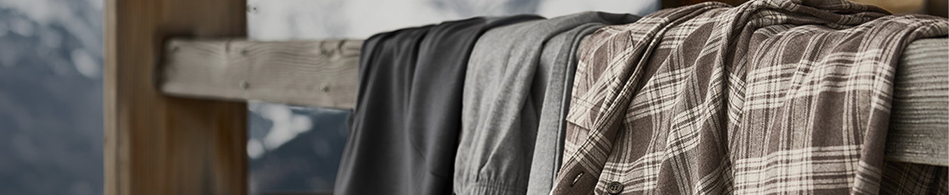 Gefaltete Herren-Loungewear und Nachtwäsche in Grau- und Beigetönen mit kariertem Muster, dekorativ über einer Holzlehne drapiert.